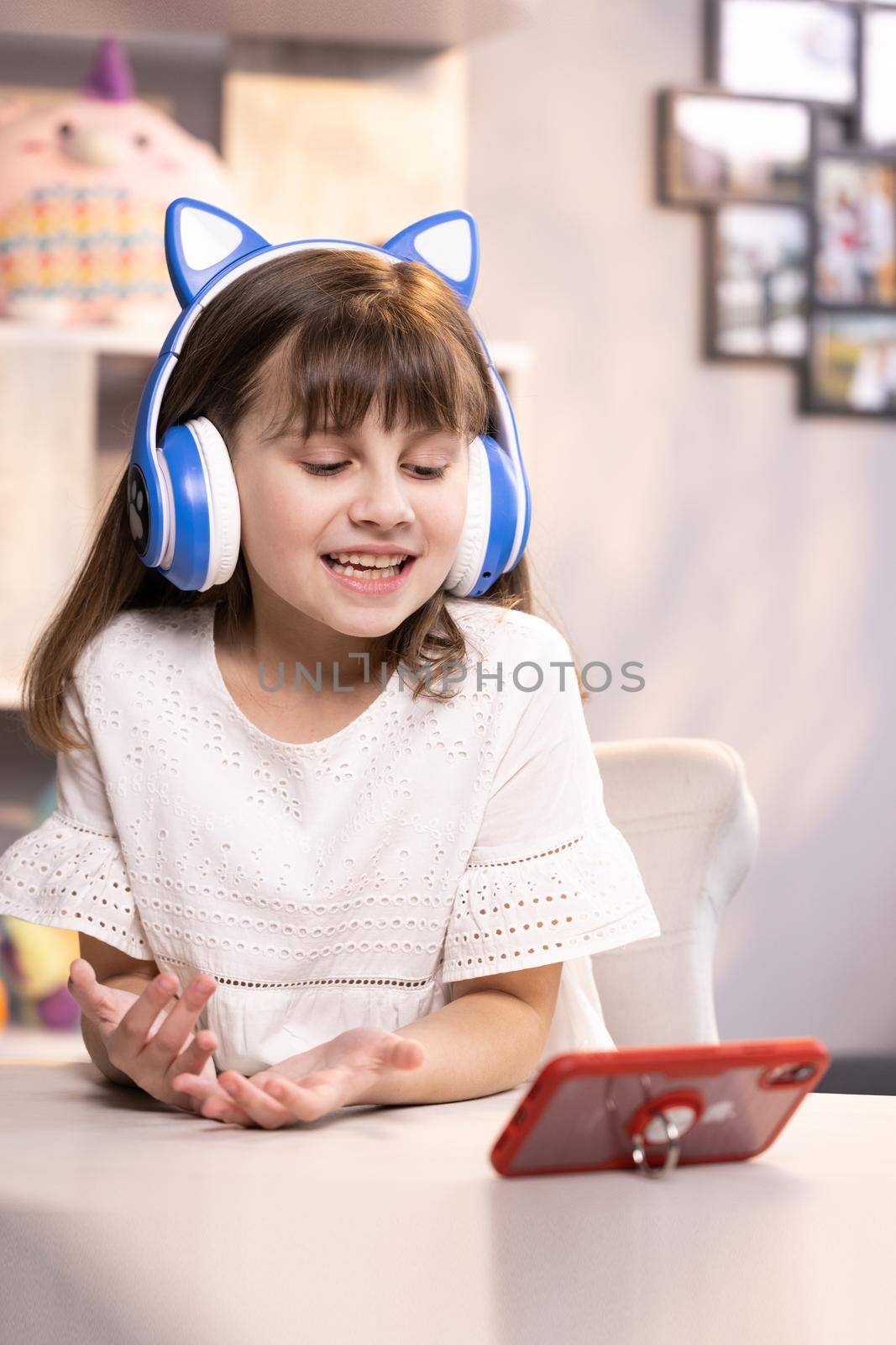 Happy cute small caucasian kid girl vlogger waving hand talking to camera at home recording vlog for social media blog, video conference calling virtual friend having online meeting sitting in chair by uflypro