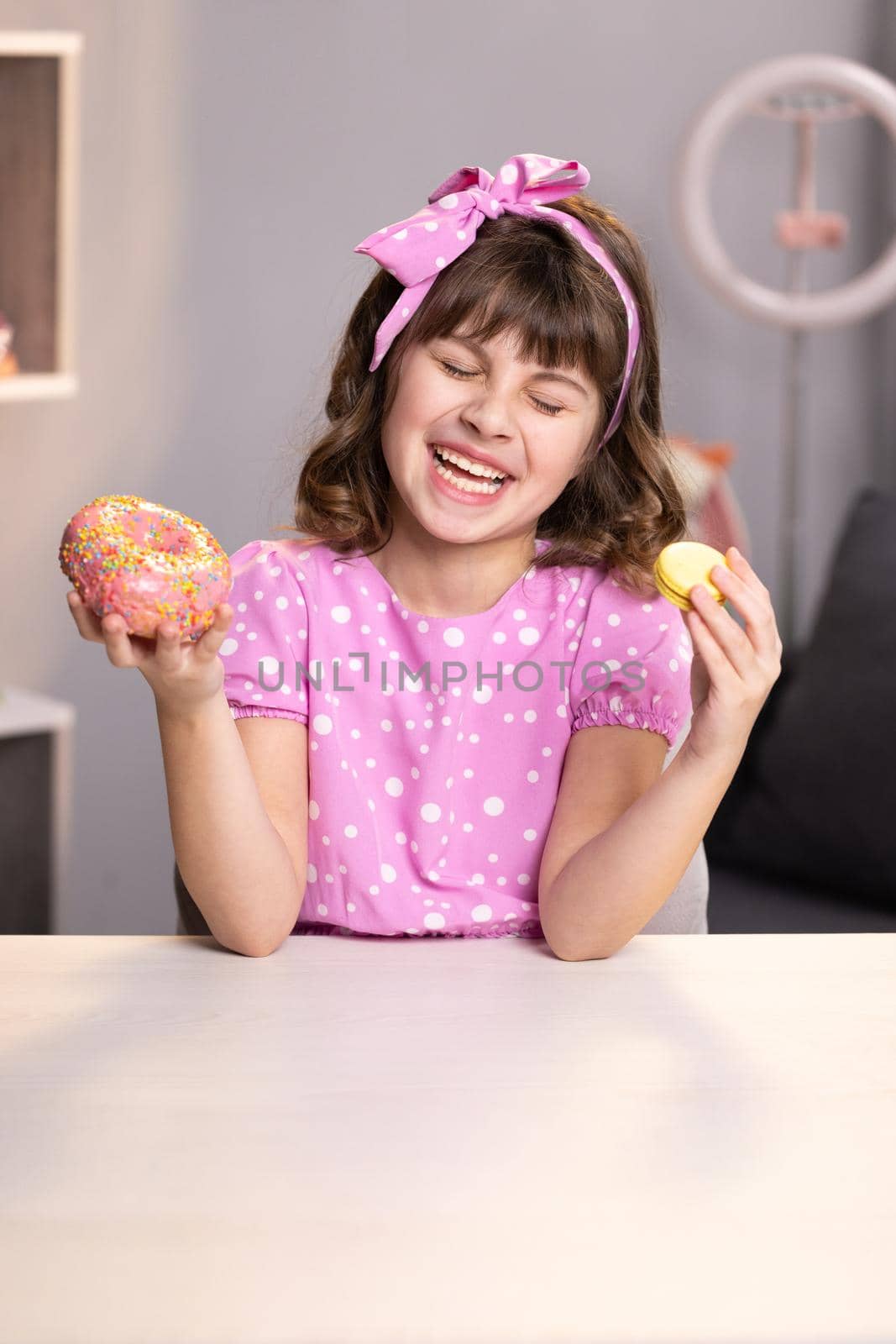 Attractive school girl sniffing donut and yellow macaron, closes her eyes with fun face and looks at camera on home background. Dessert person, sweet tooth, gourmet. Feeling good, delicious.