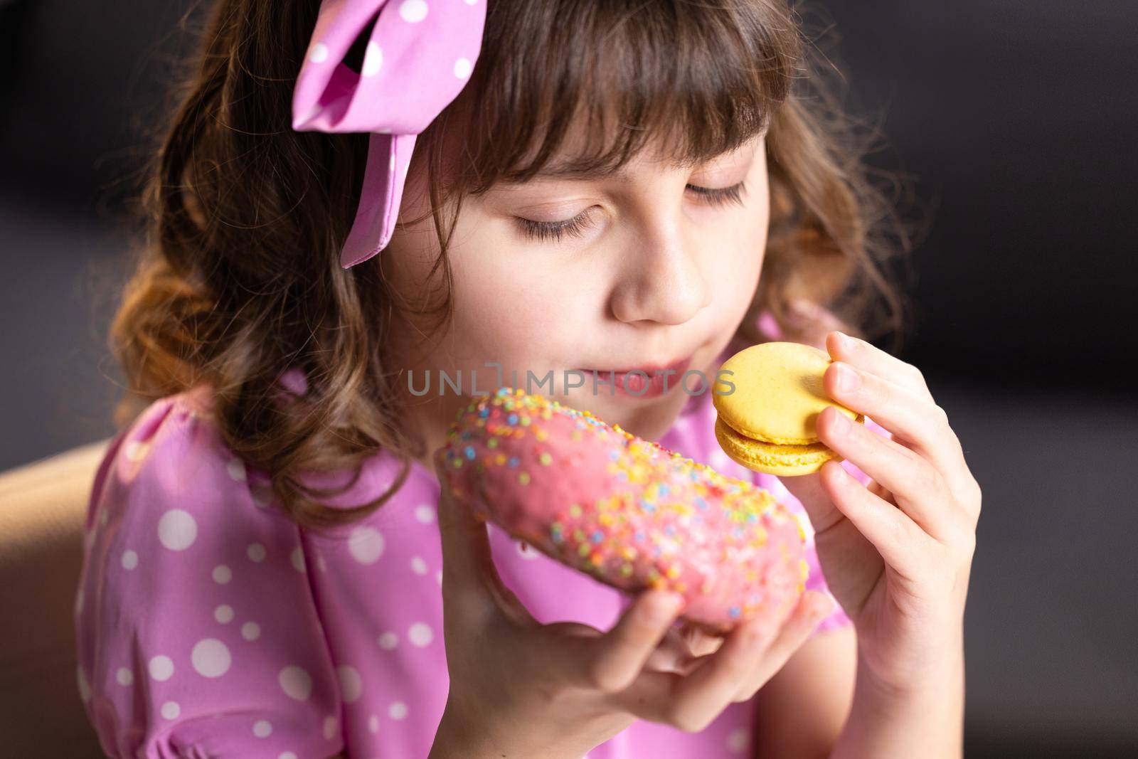 Close up of attractive school girl on home background, licks her lips, sniffing donut and yellow macaron, closes her eyes with pleasure. Dessert person, sweet tooth, gourmet. Feeling good, delicious.