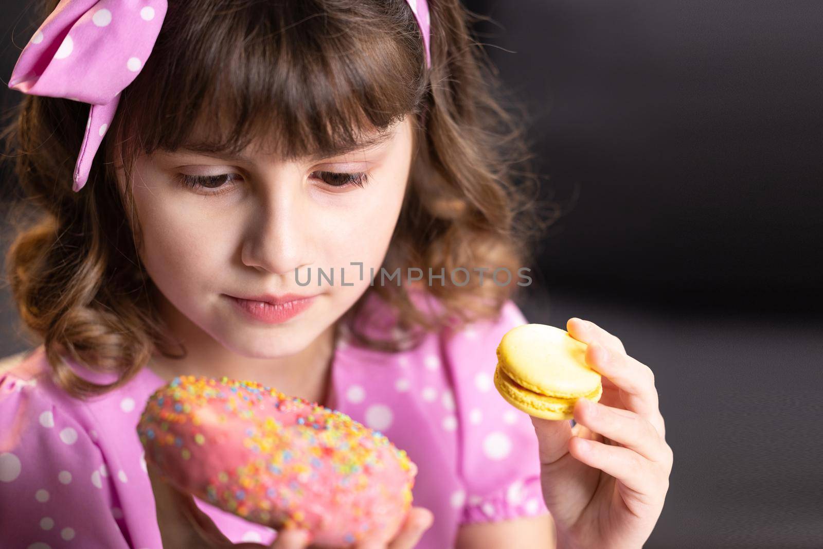 Close up of attractive school girl on home background, licks her lips, sniffing donut and yellow macaron, closes her eyes with pleasure. Dessert person, sweet tooth, gourmet. Feeling good, delicious.