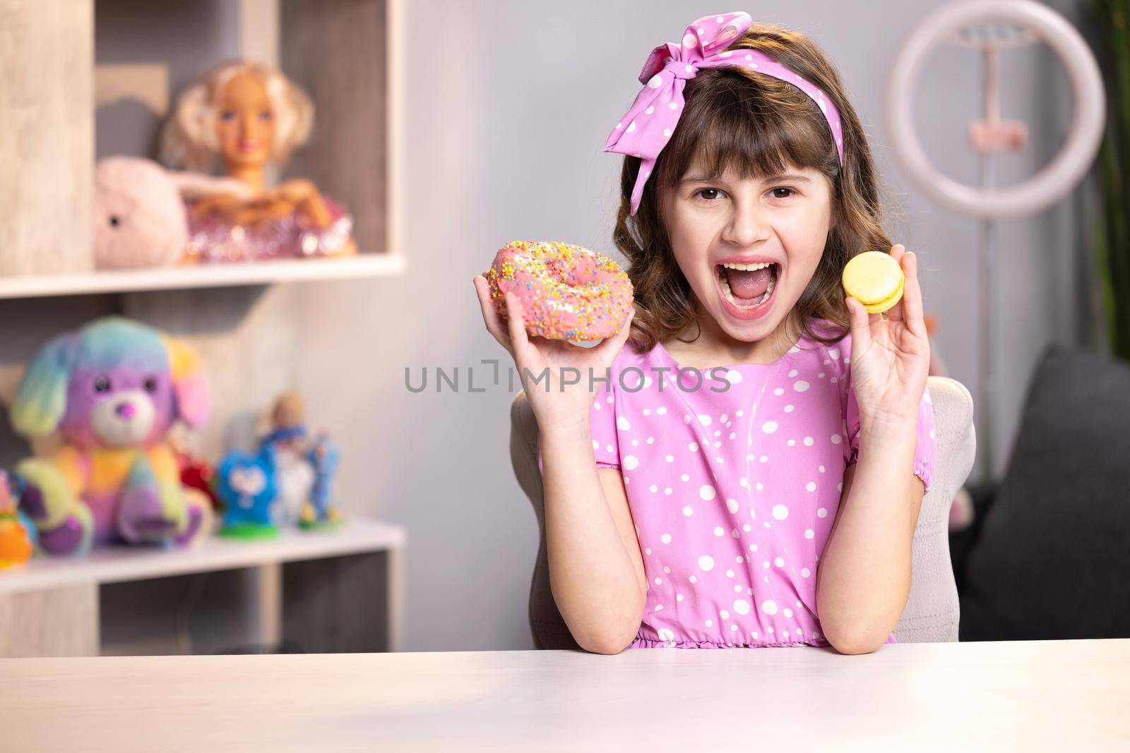 Cute little girl in pink dress holding macaron and donut in hands by the face. Adolescent school girl plays with sweet donuts doing happy fun face expressions on background. Funny concept with sweets.