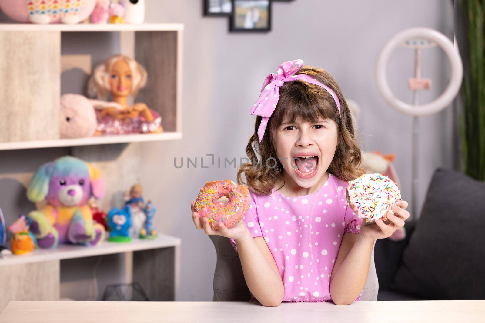 Cute adolescent school girl plays with sweet donuts doing happy fun face expressions on background. Little girl in pink dress holding two donuts in hands by the face. Funny concept with sweets.