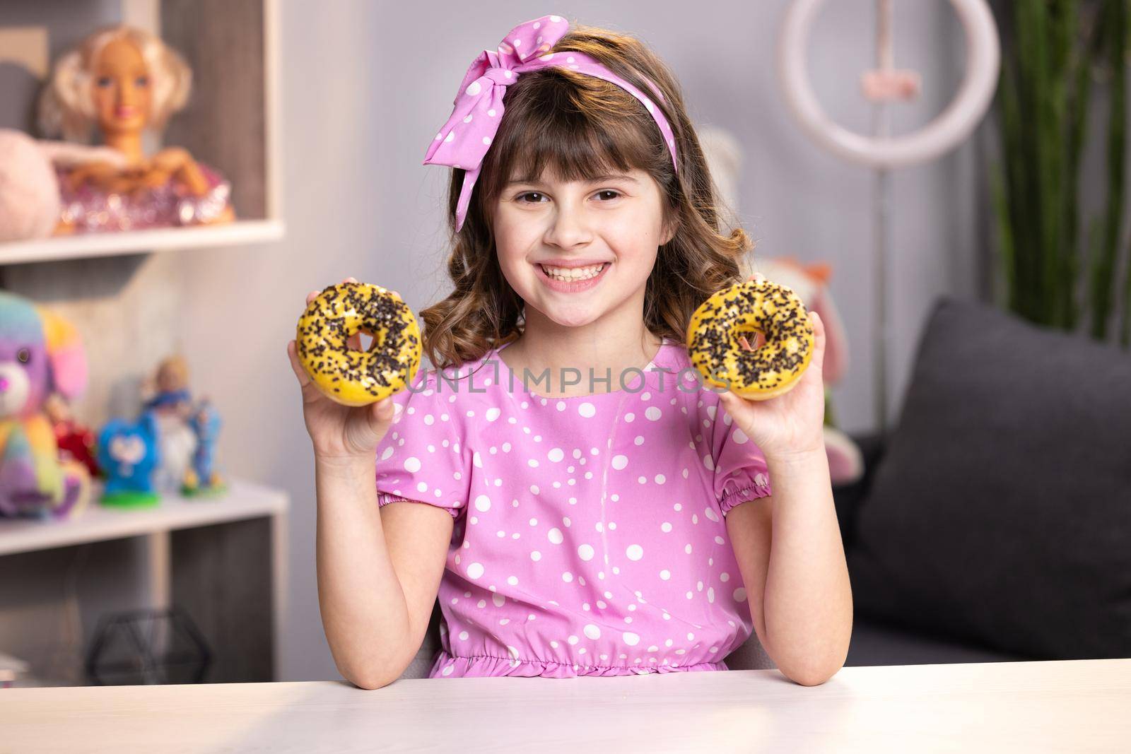 Portrait of sweet girl choosing between two donuts in home room. Cheerful school girl playing with cakes indoors. Funny teenager girl having fun with colorful donuts at modern home.