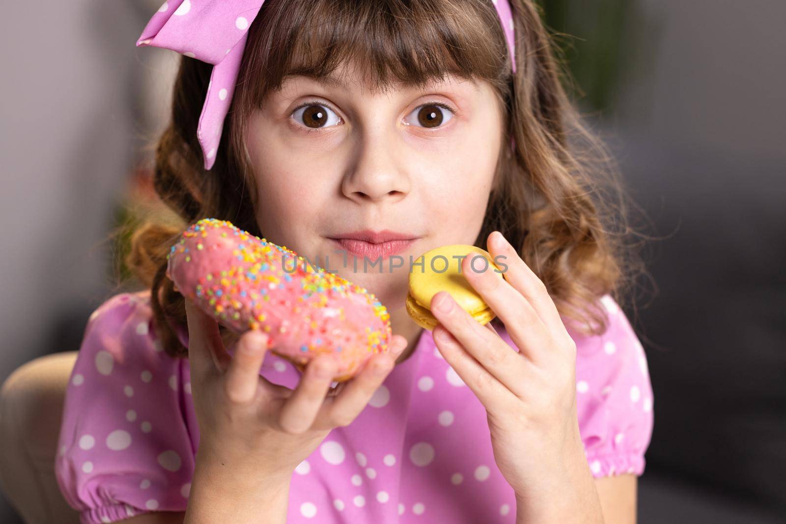 Cute little girl in pink dress holding macaron and donut in hands by the face. Funny concept with sweets. Adolescent school girl plays with sweet donuts doing happy fun face expressions on background.