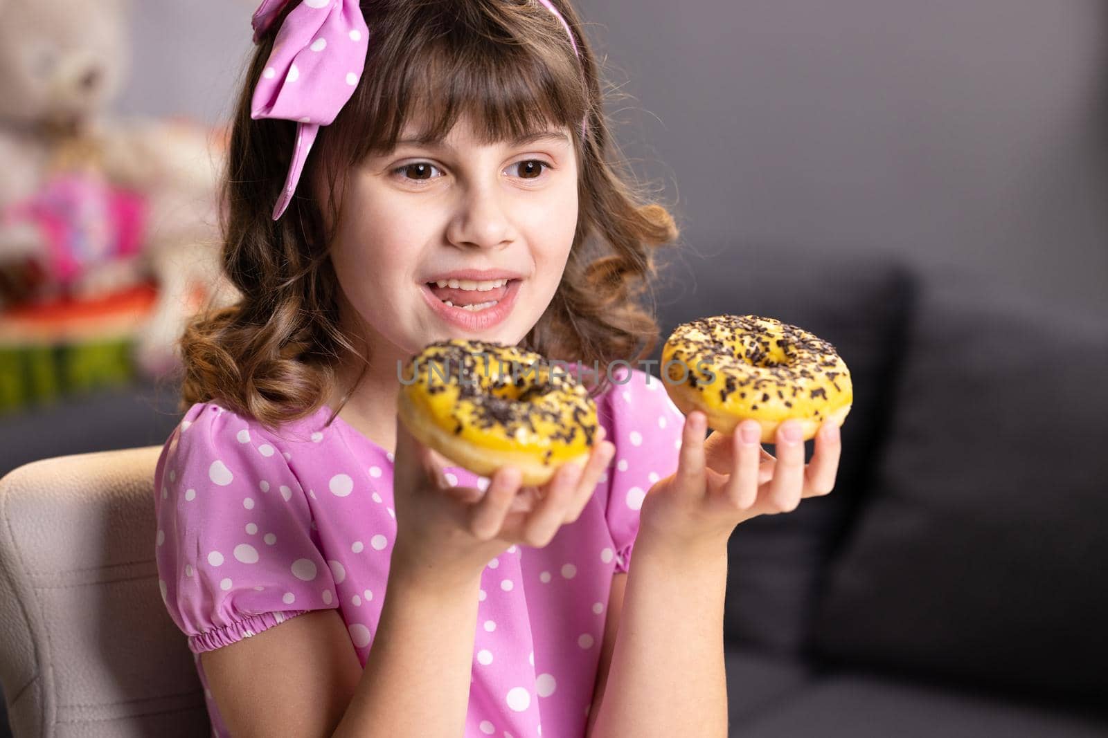 Cheerful school girl playing with cakes indoors. Funny teenager girl having fun with colorful donuts at modern home. Portrait of sweet girl choosing between two donuts in home room
