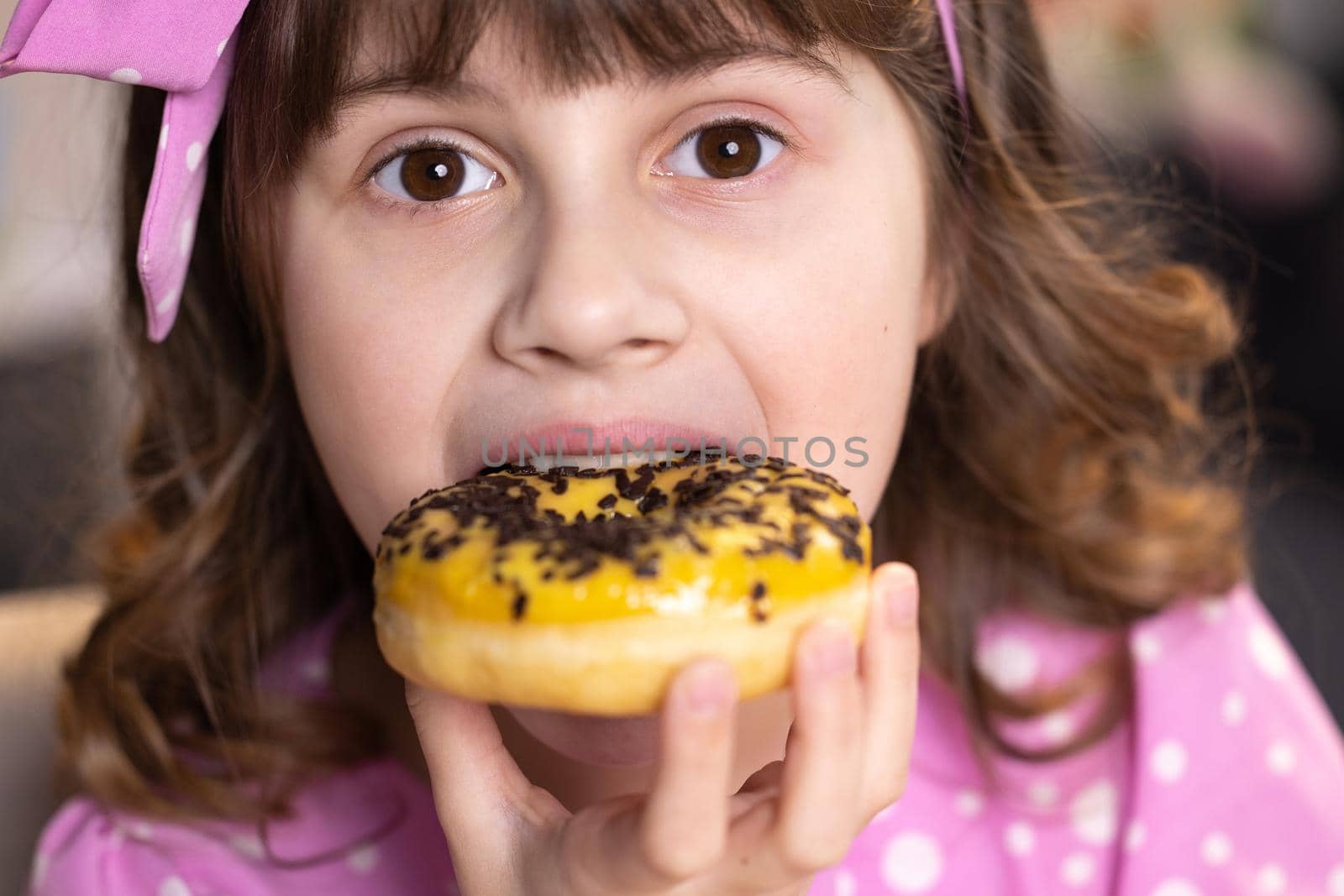Close up school girl eating donut while sitting at table at home. Portrait of sweet girl choosing donut indoors. Funny teenager girl having fun with yellow donuts at modern home.