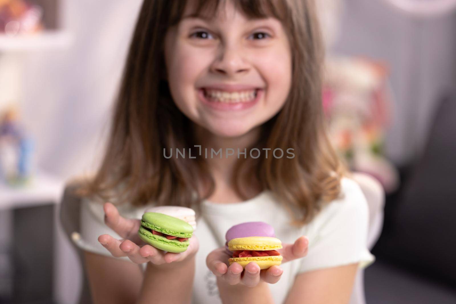 Smiling cute school kid girl holds four tasty cookies in her hands and look at camera. Traditional French multicolored macaroon. Food concept by uflypro