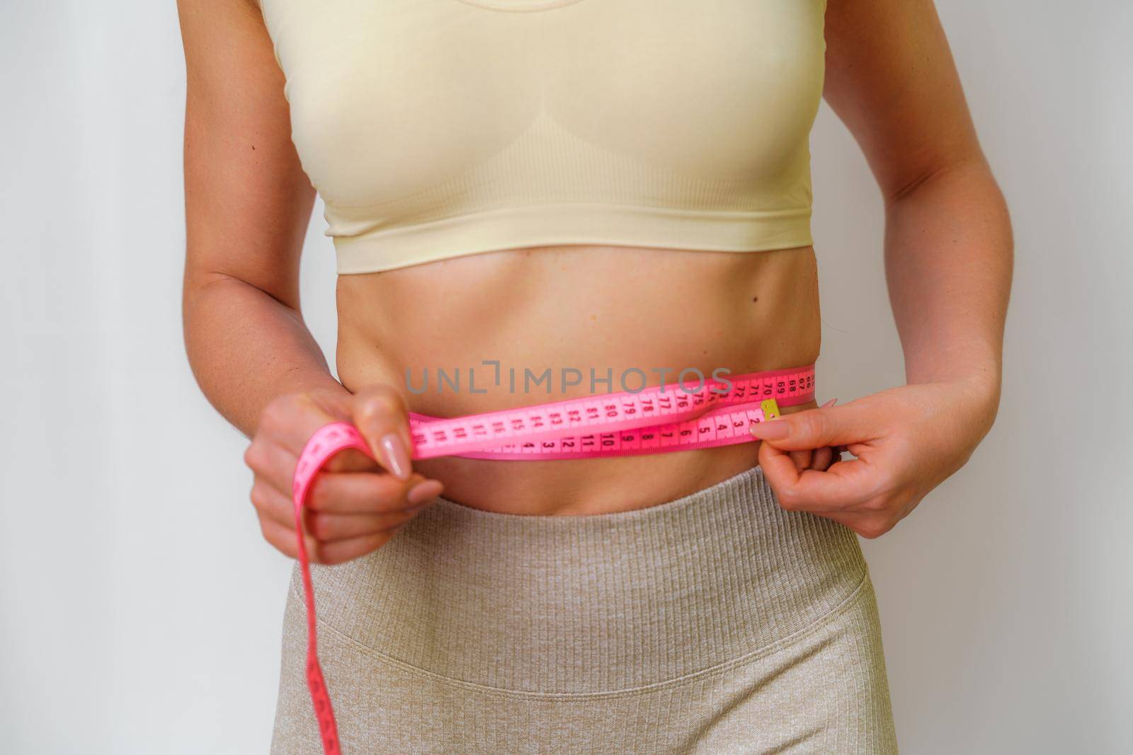 Cropped view of slim woman measuring waist with tape measure at home, close up. An unrecognizable European woman checks the result of a weight loss diet or liposuction indoors. Healthy lifestyle. by Matiunina