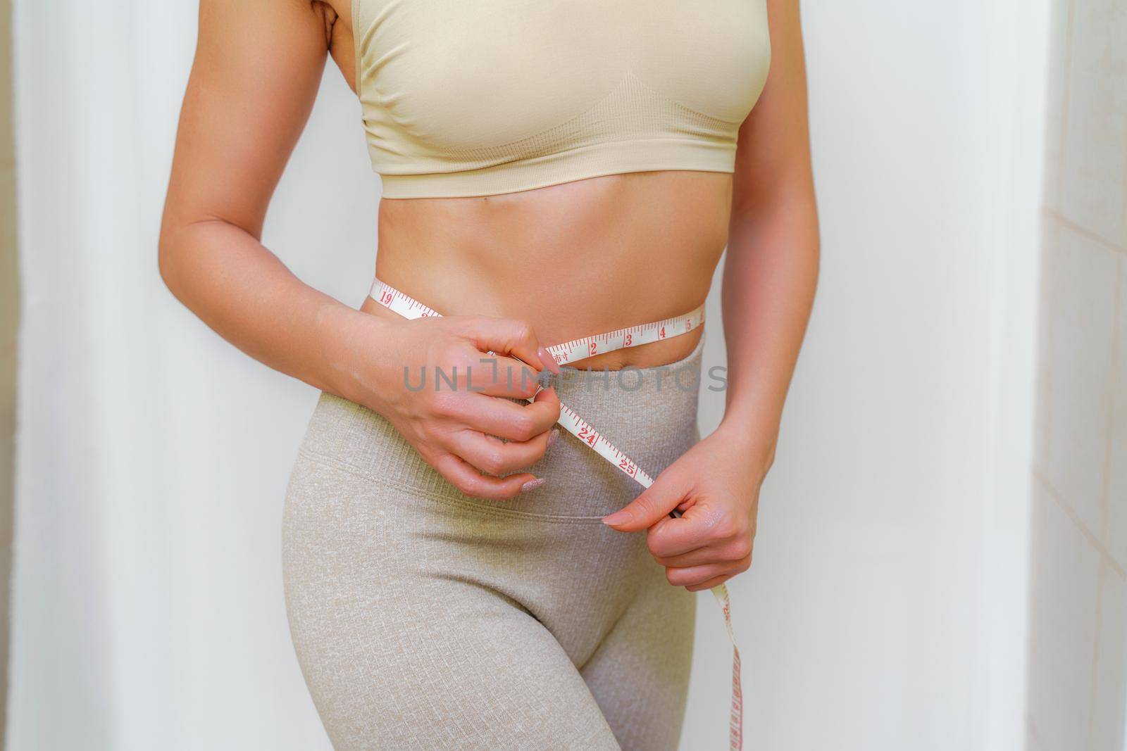Cropped view of slim woman measuring waist with tape measure at home, close up. An unrecognizable European woman checks the result of a weight loss diet or liposuction indoors. Healthy lifestyle. by Matiunina