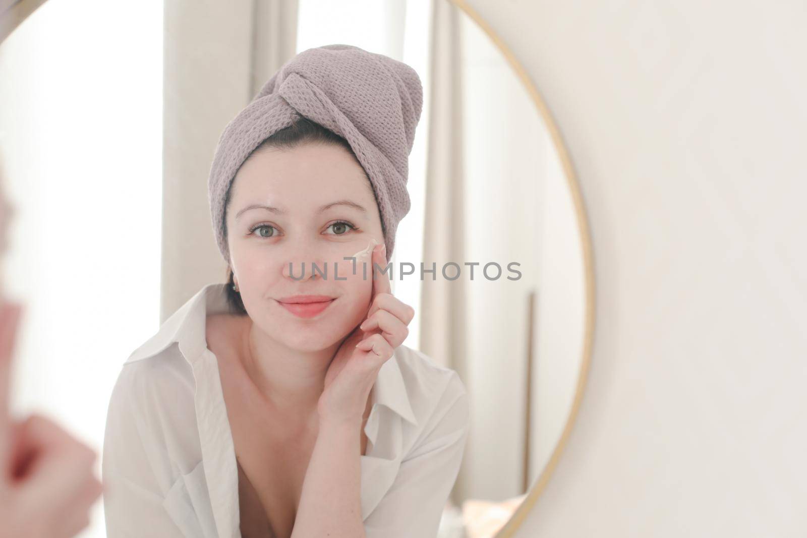 young woman with a towel on head looking in the mirror and apply cream on face skin.