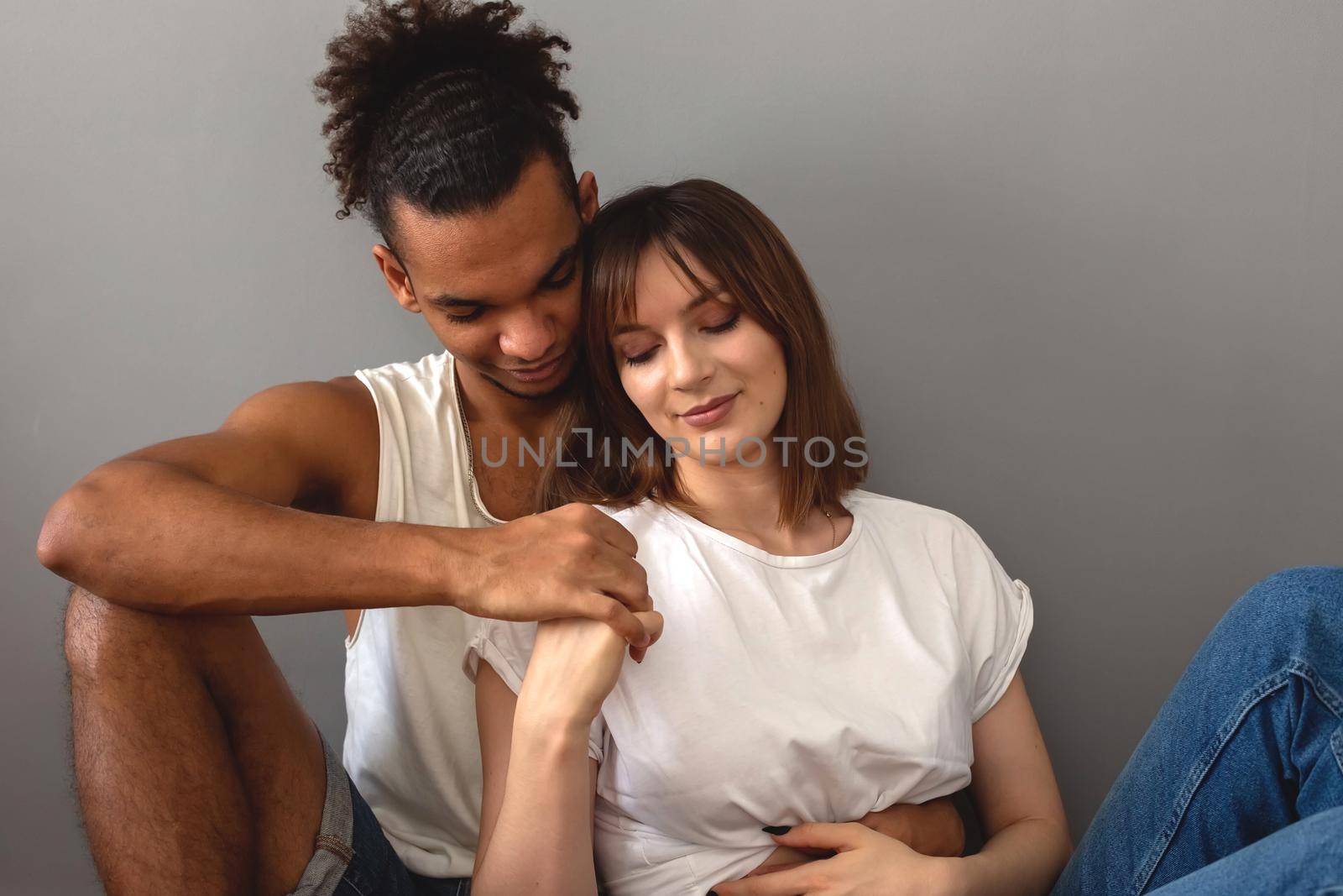 Multiracial family, guy and girl in white t-shirts are sitting at home on the floor against a gray wall, hugging, gentle touches.