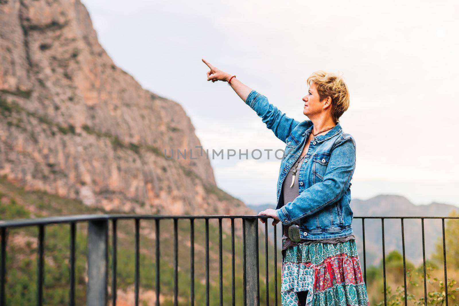 Adult woman, blonde, short hair, looking and pointing at a mountain view from a lookout point, on holiday. by CatPhotography