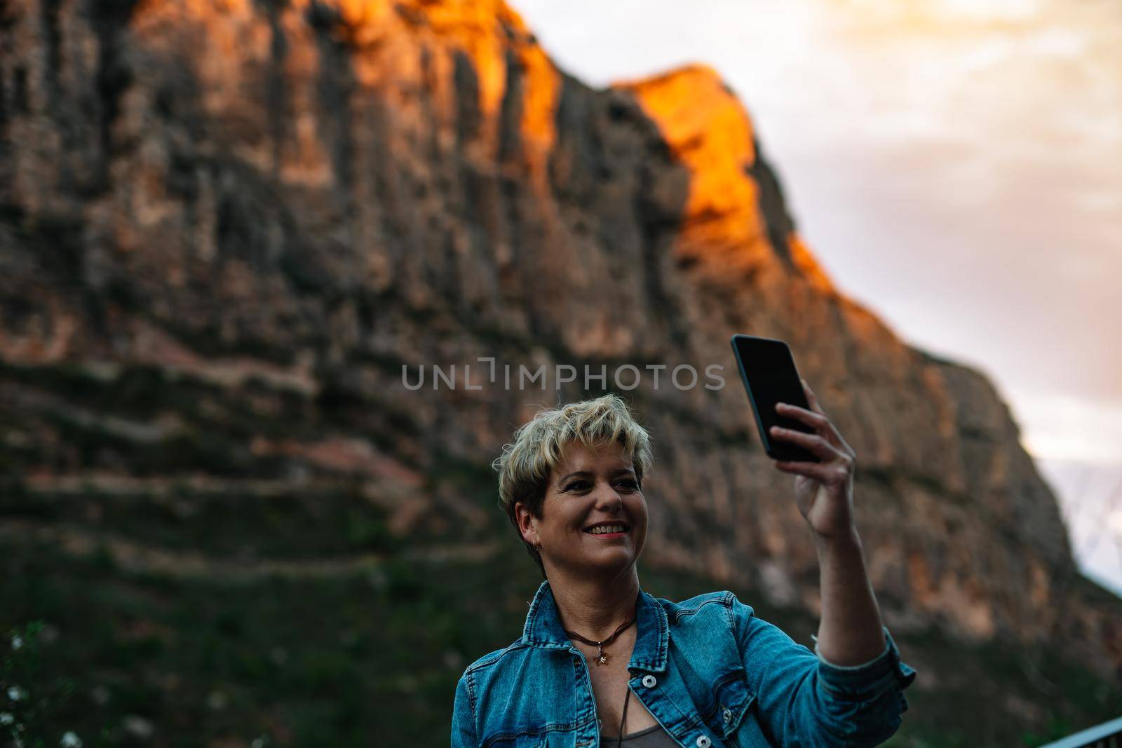 Portrait of an adult blonde short haired modern woman, smiling and happy, casually dressed, wearing a blue denim jacket, taking a selfie on holiday. Warm and calm atmosphere with sunset light, background of mountains and forests, blue sky with big clouds.