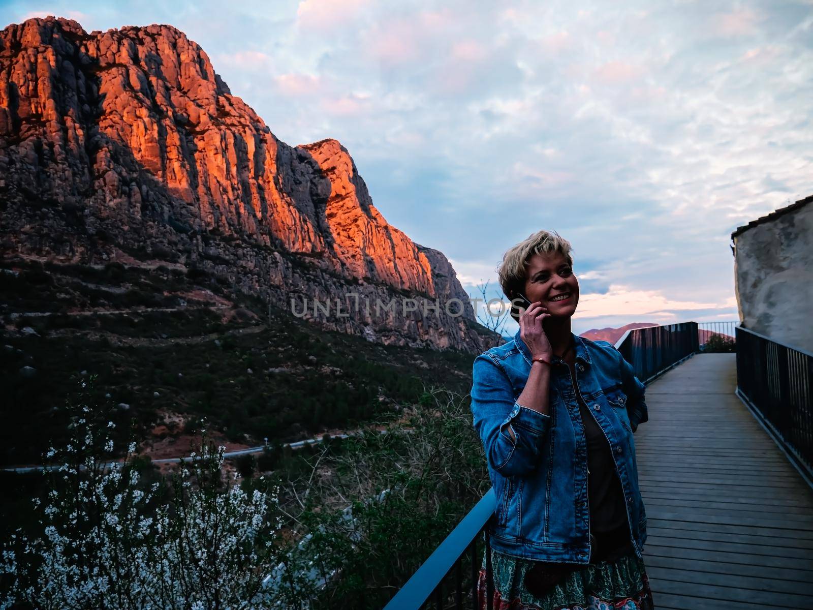 Portrait of an adult blonde short haired modern woman, smiling and happy, casually dressed, wearing blue denim jacket, long coloured skirt, on holiday walking and talking on the phone with her family, tells how she enjoys her getaway. Warm and calm atmosphere with sunset light, background of mountains and forests, blue sky with big clouds.