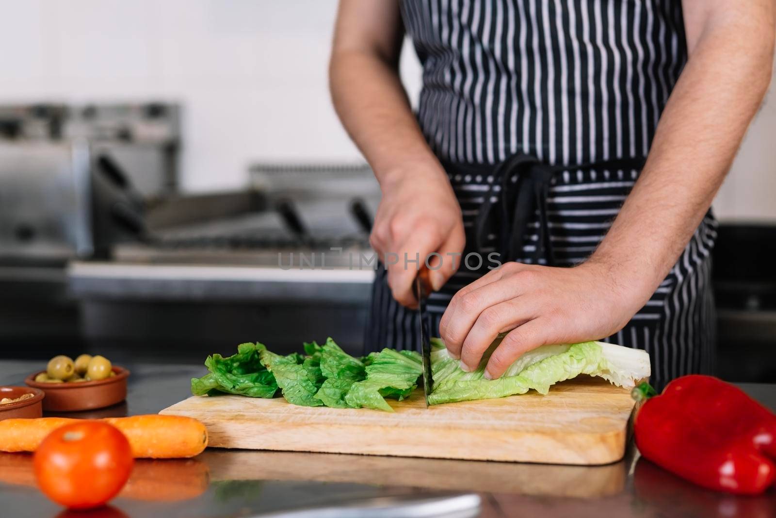 detail of the hands of a young man, dressed in a black polo shirt and a dark striped apron, cutting vegetables with a knife on a wooden board in a professional kitchen, on a table full of food. Cold and clean environment, background of professional aluminium tables and cooking fires of a restaurant.