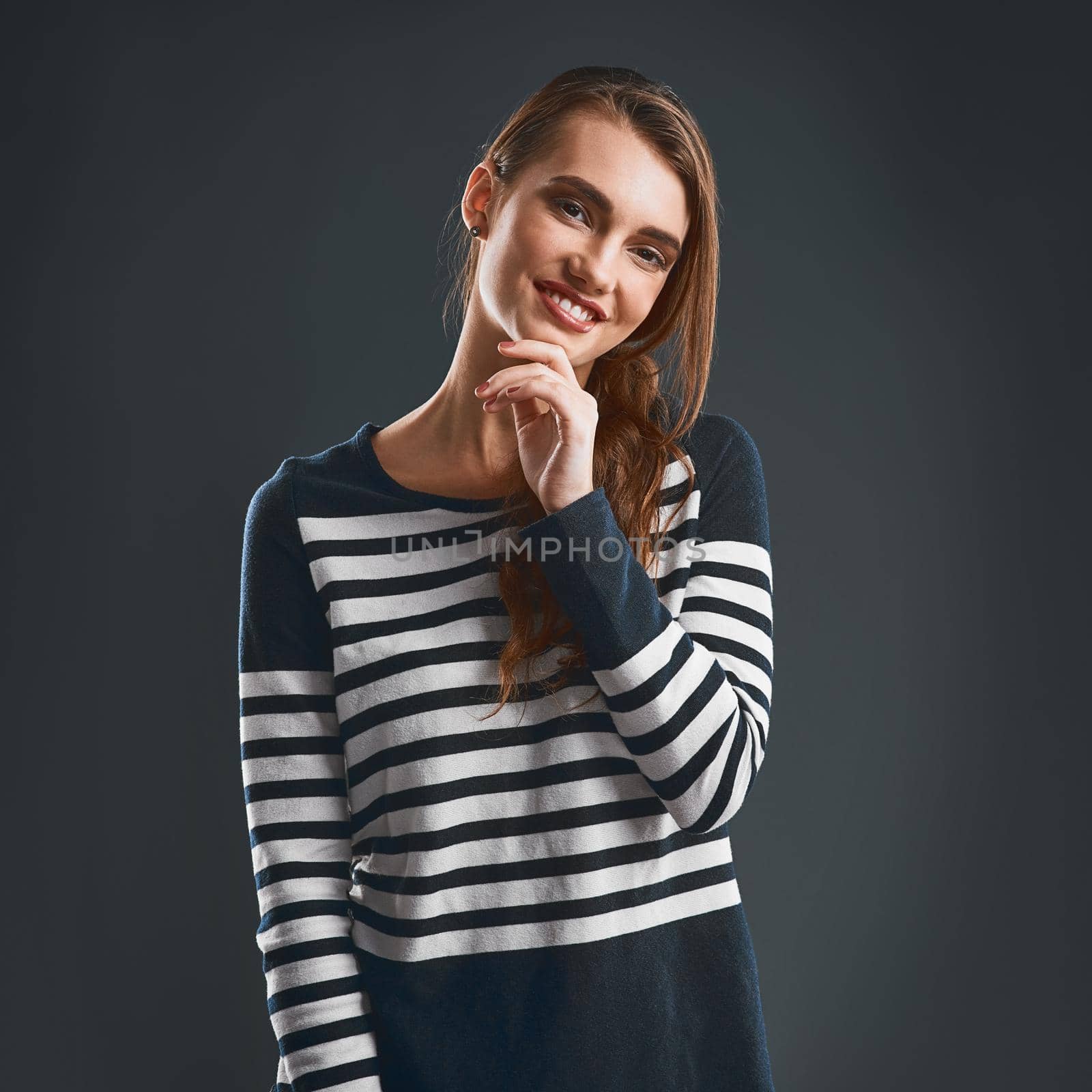 Studio portrait of a cheerful young woman holding her chin while standing against a dark background.