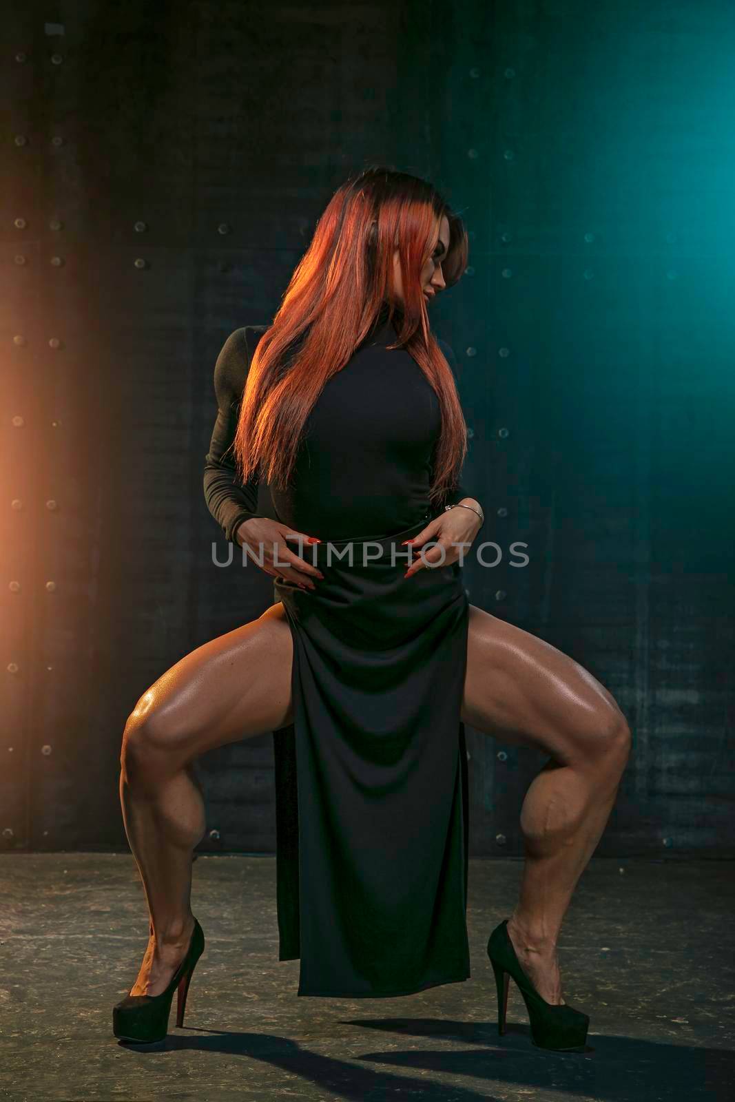 Graceful red-haired girl dances in black dress in the studio in the spotlight on black background.