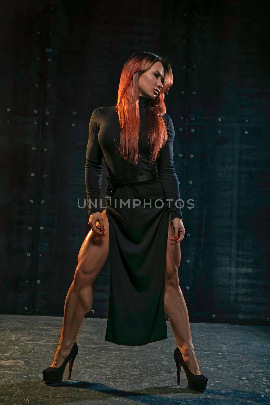 Graceful red-haired girl in black dress with high heels in the studio on metallic background