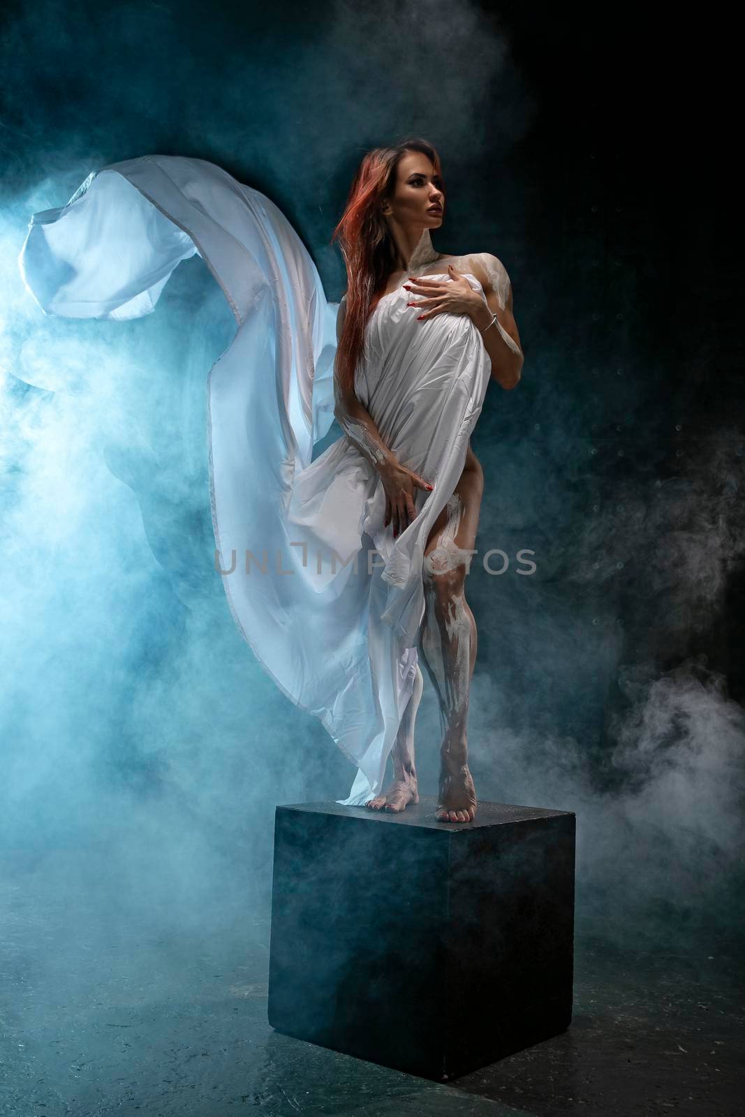 Red-haired girl covers herself with white cloth like an antique statue, posing in the studio by but_photo