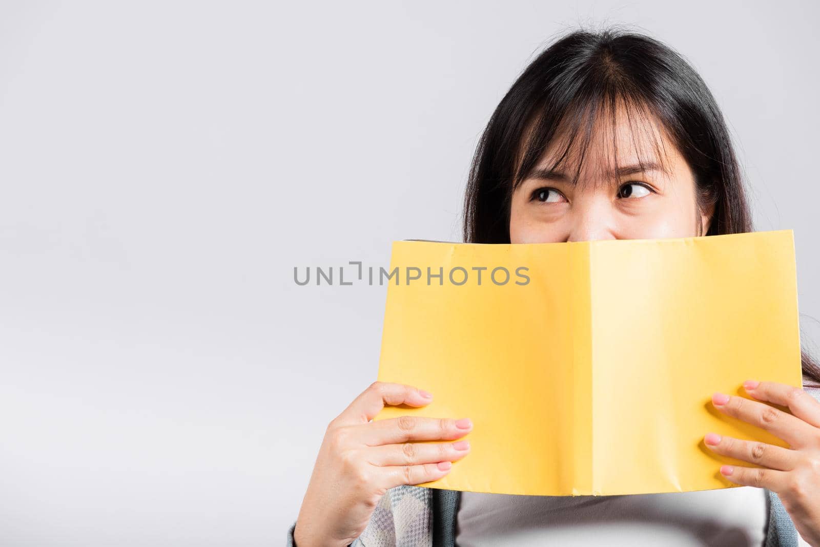 Woman teen smiling covering her face with open book, Portrait of beautiful Asian young female person hiding behind an book show only eyes studio shot isolated on white background, education concept