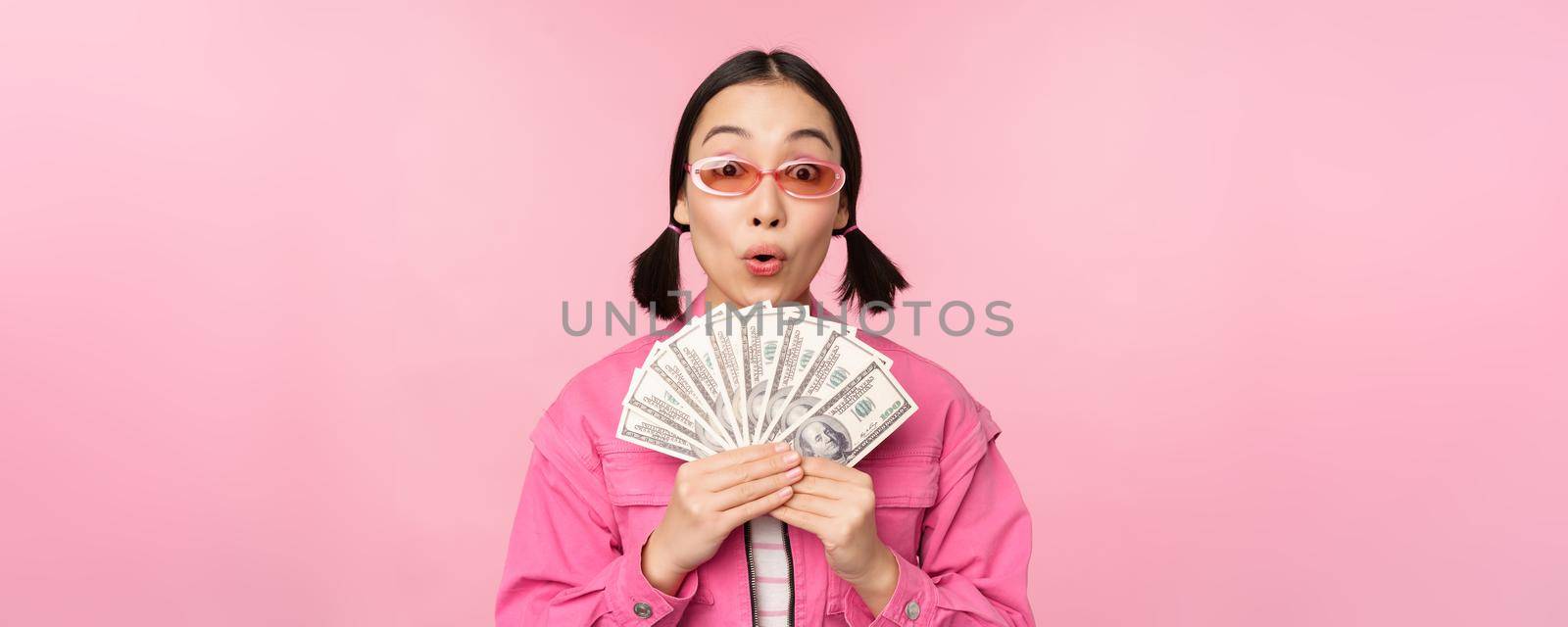 Microcredit and fast loans concept. Excited stylish korean girl, showing money, cash dollars and looking happy, standing in sunglasses over pink background by Benzoix