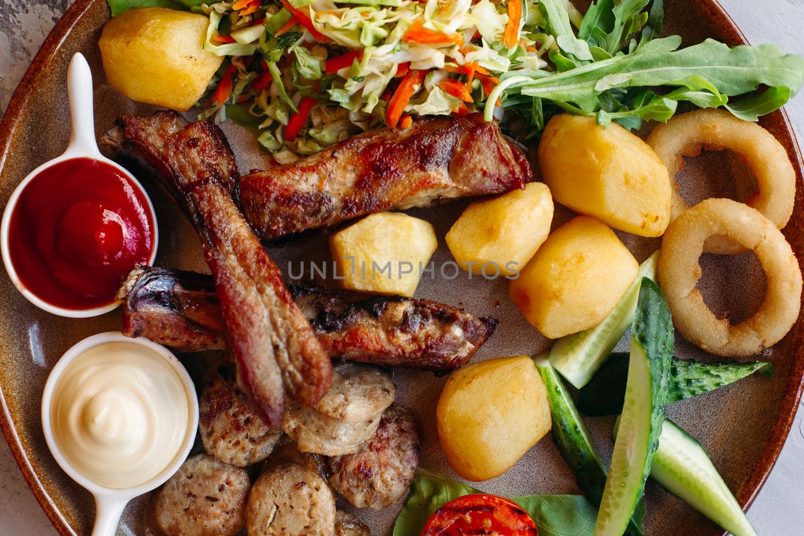 Horizontal photo of tasty food laying on clay plate. Delicious set of vegetable salad, boiled potatoes, grilled chicken legs, roasted onion, sausages, cucmbers, tomatoes and sauces - ketchup and mayo.