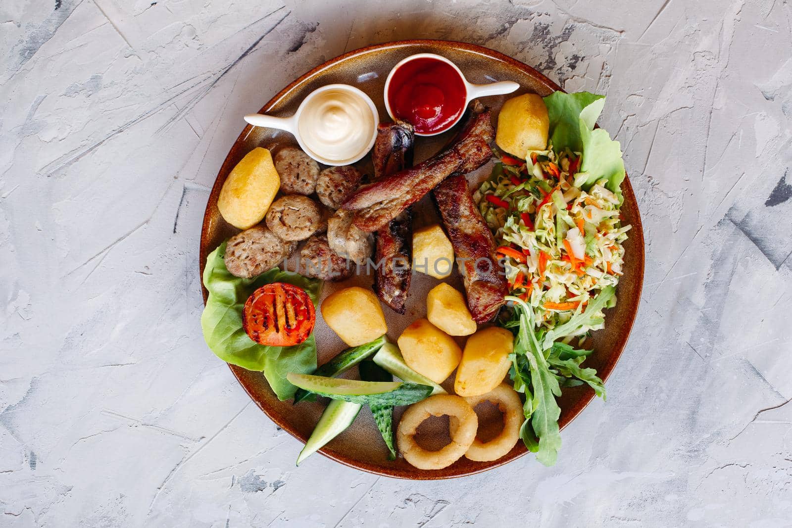 Top view of clay plate full of appetizers for beer including vegetable salad, boiled potatoes, grilled chicken legs, roasted goldy onion and sausages, cucmbers, tomatoes and sauces - ketchup and mayo.