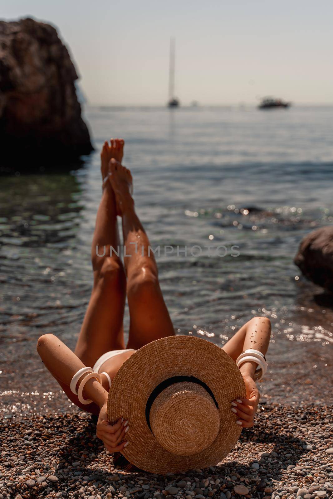 A beautiful middle-aged woman lies on the beach with her feet to the sky, covering her body with a wide-brimmed straw hat. She is sunbathing on the ocean. Vacation, travel, vacation concept