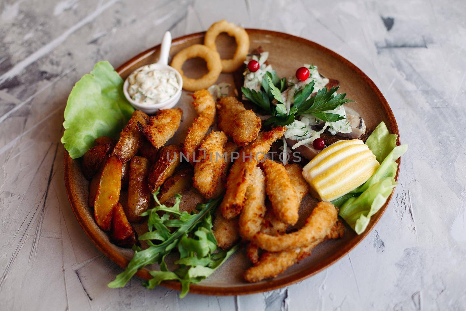 Clay plate full of appetizers including goldy chicken nuggets with chrispy crust, delicious canapes with herring and cherry tomatoes, served with garlic sauce, decorated with salad leaves and cheese.