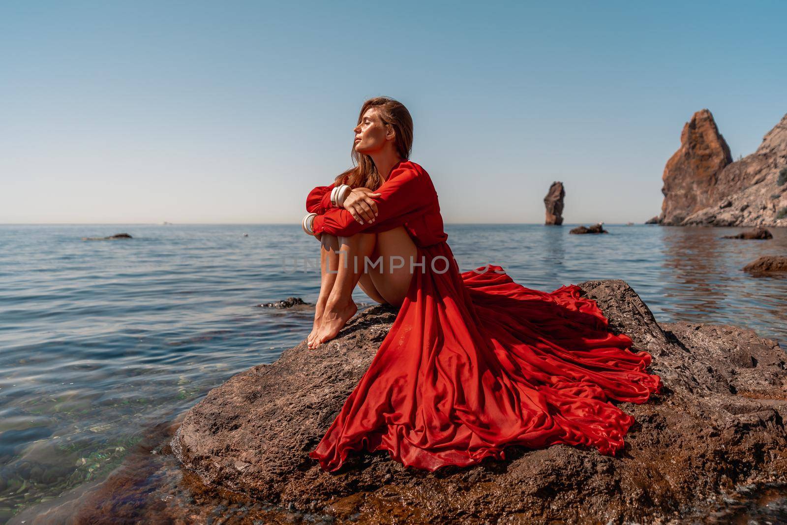 Beautiful sensual woman in a flying red dress and long hair, sitting on a rock above the beautiful sea in a large bay. by Matiunina
