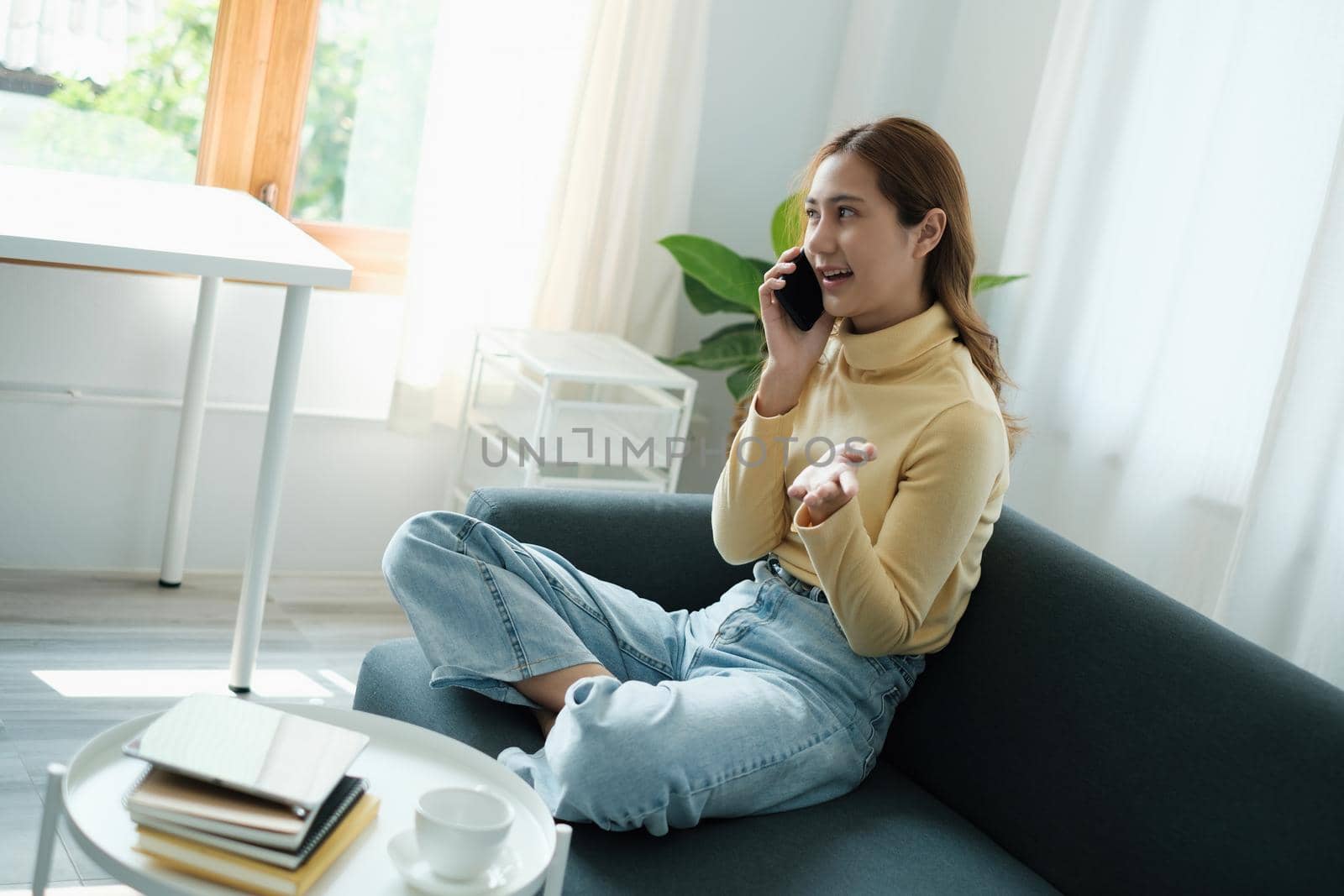 Young Asian woman talking with friend by cell phone while sitting on sofa at home