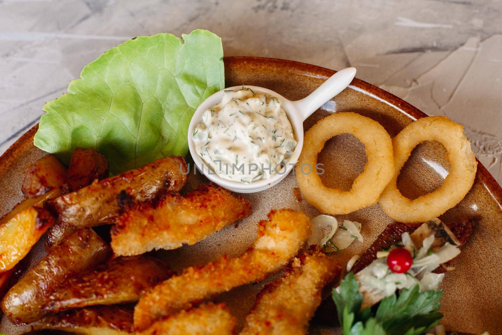 Delicious appetizers for beer and light alcohol drinks laying on clay plate decorated with salad leaves and cheese: crispy chicken nuggets, canapes with herring and cherry tomatoes, garlic sauce.