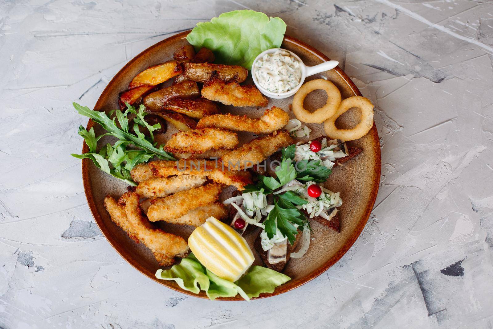 Clay plate full of appetizers including goldy chicken nuggets with chrispy crust, delicious canapes with herring and cherry tomatoes, served with garlic sauce, decorated with salad leaves and cheese.