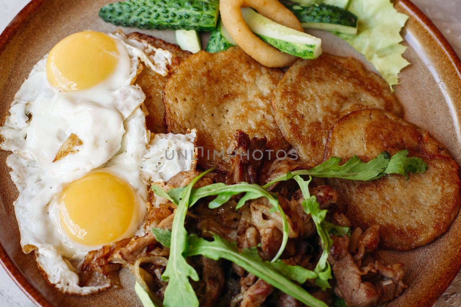 Delicious potato puncakes lying on brown clay plate with fried eggs. Fresh green cucumbers, fried golden onion and crusty meat, served with salad leaves. Tasty mouthwatering morning's meal.