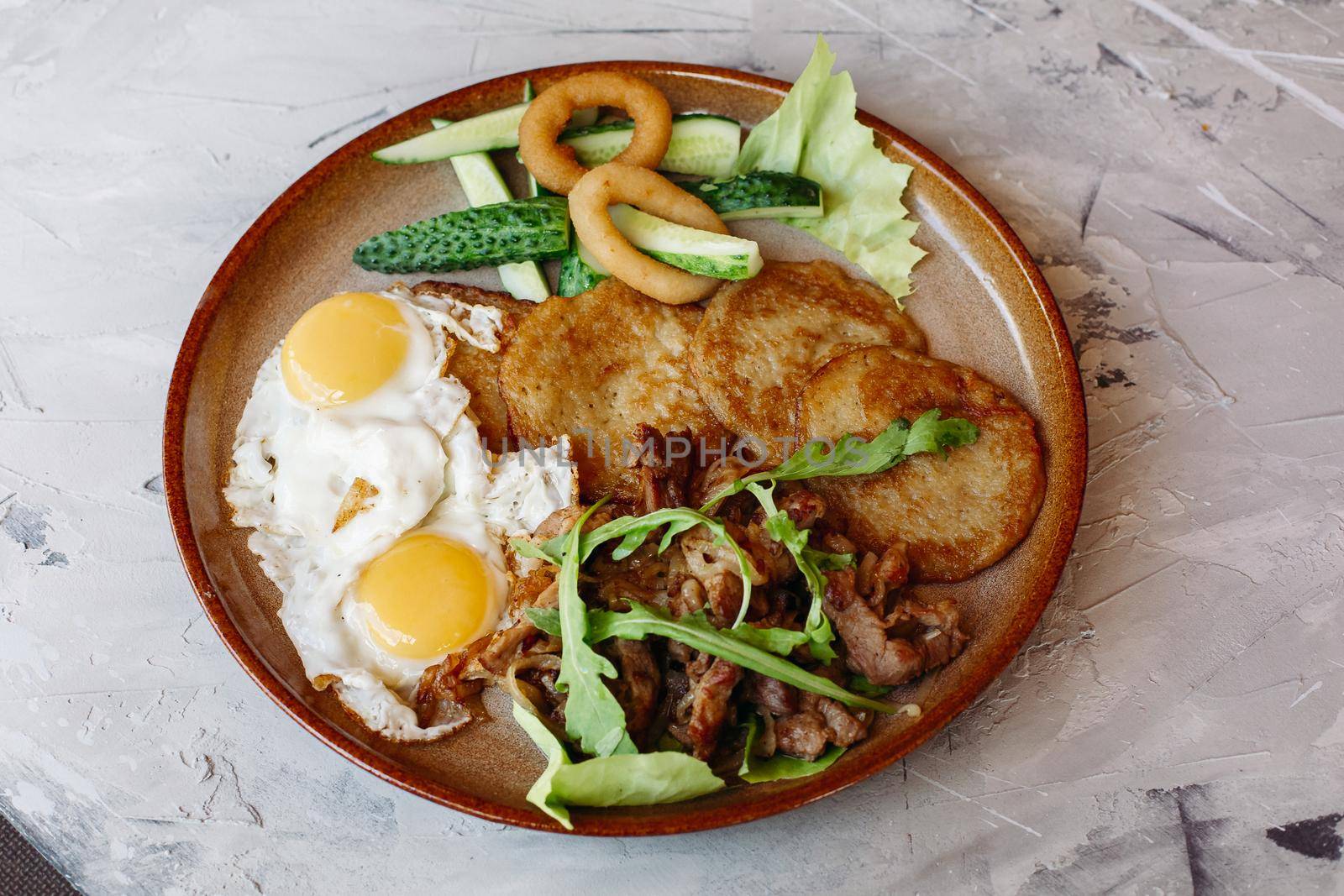 Delicious potato puncakes served with fried eggs laying on glancy clay plate including fresh green cucumbers, fried golden onion and crusty meat, served with salad leaves. Looking tasty. White table.