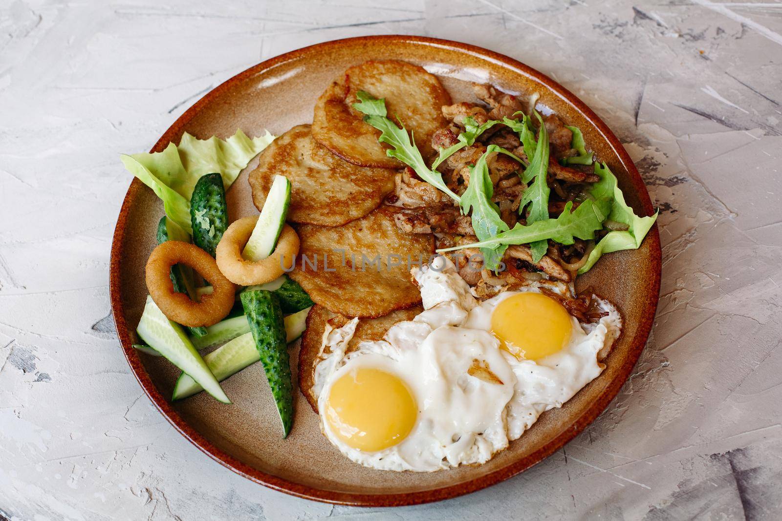 Delicious potato puncakes served with fried eggs laying on glancy clay plate including fresh green cucumbers, fried golden onion and crusty meat, served with salad leaves. Looking tasty. White table.