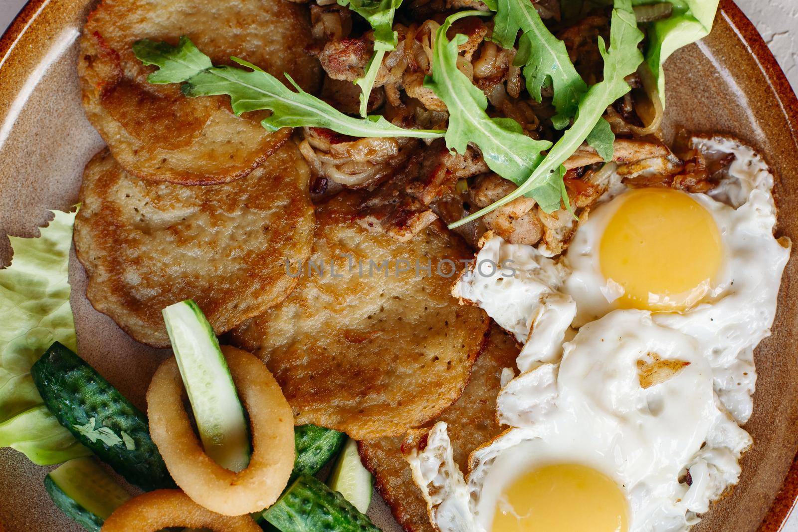 Delicious potato puncakes lying on brown clay plate with fried eggs. Fresh green cucumbers, fried golden onion and crusty meat, served with salad leaves. Tasty mouthwatering morning's meal.