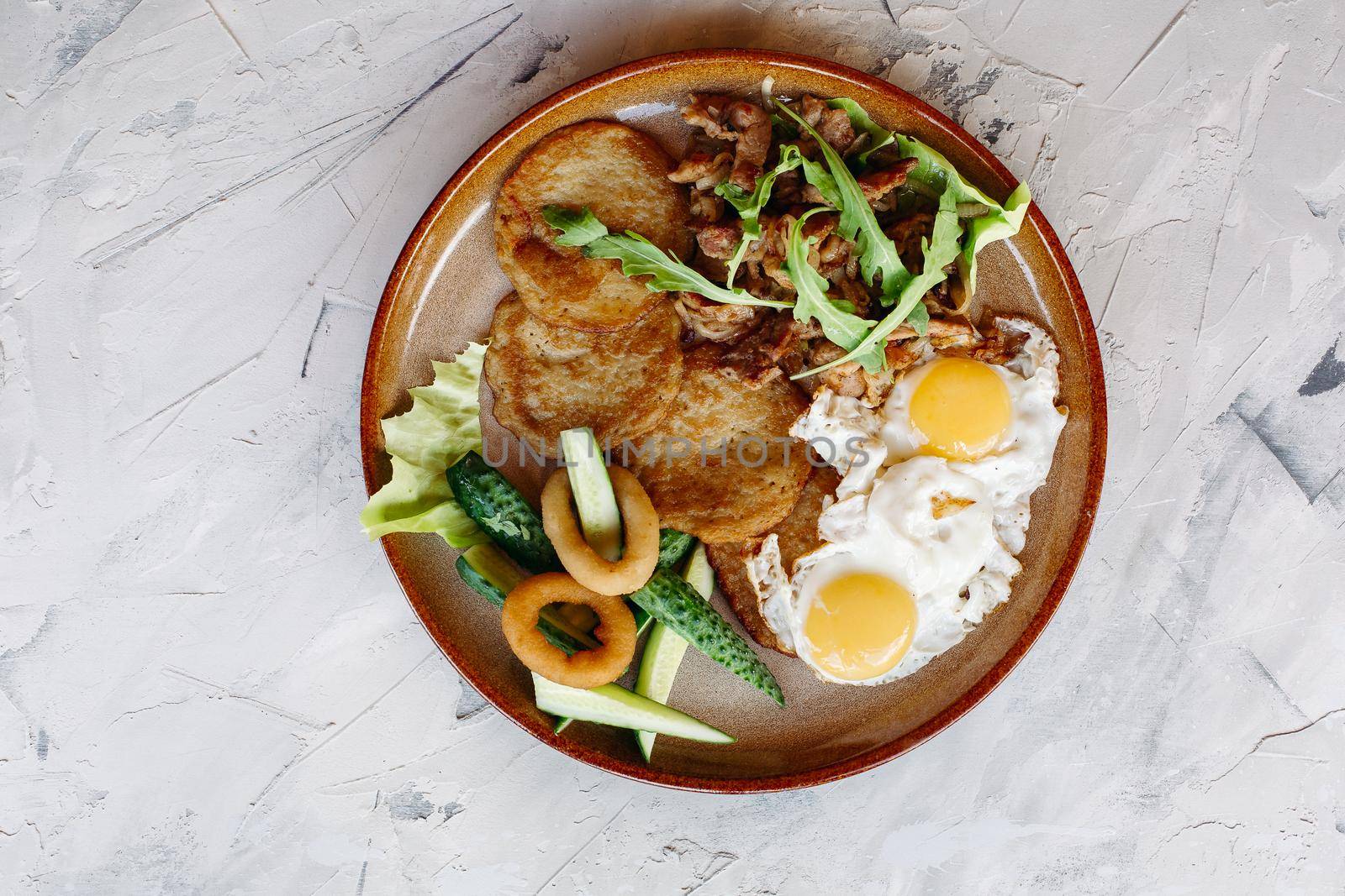 Delicious potato puncakes served with fried eggs laying on glancy clay plate including fresh green cucumbers, fried golden onion and crusty meat, served with salad leaves. Looking tasty. White table.