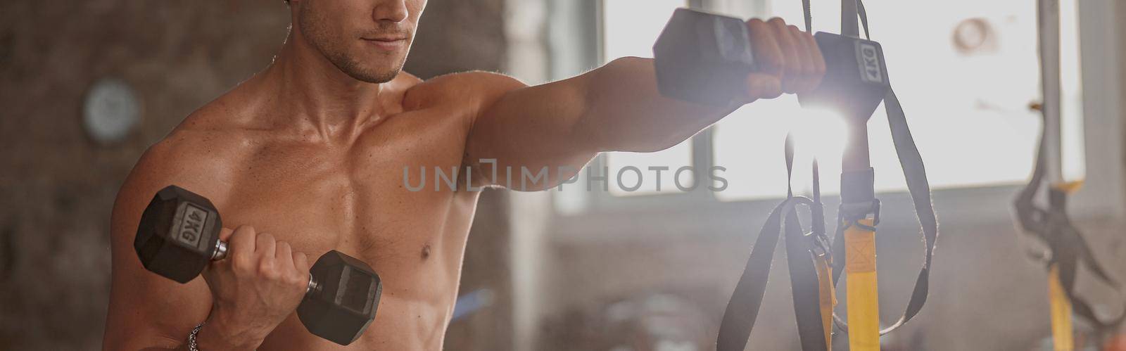 Muscular young man with beautiful athletic body doing bodybuilding exercises with dumbbells in gym