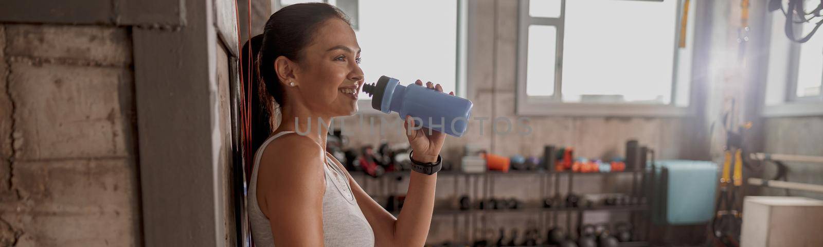 Happy lady leaning against wall at the sport club, hand on thigh, drinking water looking aside
