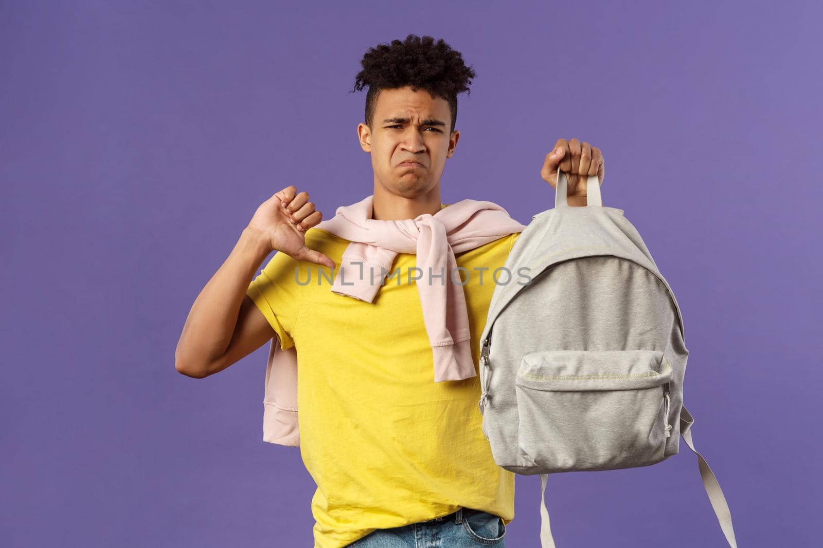 Education, university and trends concept. Portrait of gloomy disappointed young male student complaining on ugly new backpack, show thumbs-down and grimacing displeased, purple background.