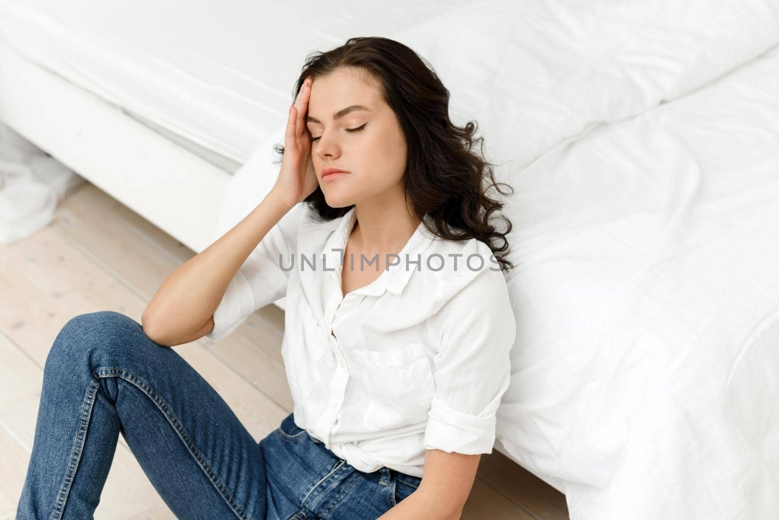Unhappy woman touches her head, sits on the floor by the bed at home, she has a headache. Young frustrated woman thinks about problems, feels lonely and sad.