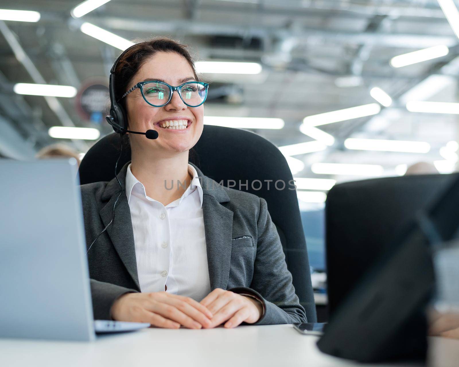 A happy female support operator is sitting at a desk and answering calls. Beautiful smiling woman talking to customers on a headset. Office employee in headphones. by mrwed54