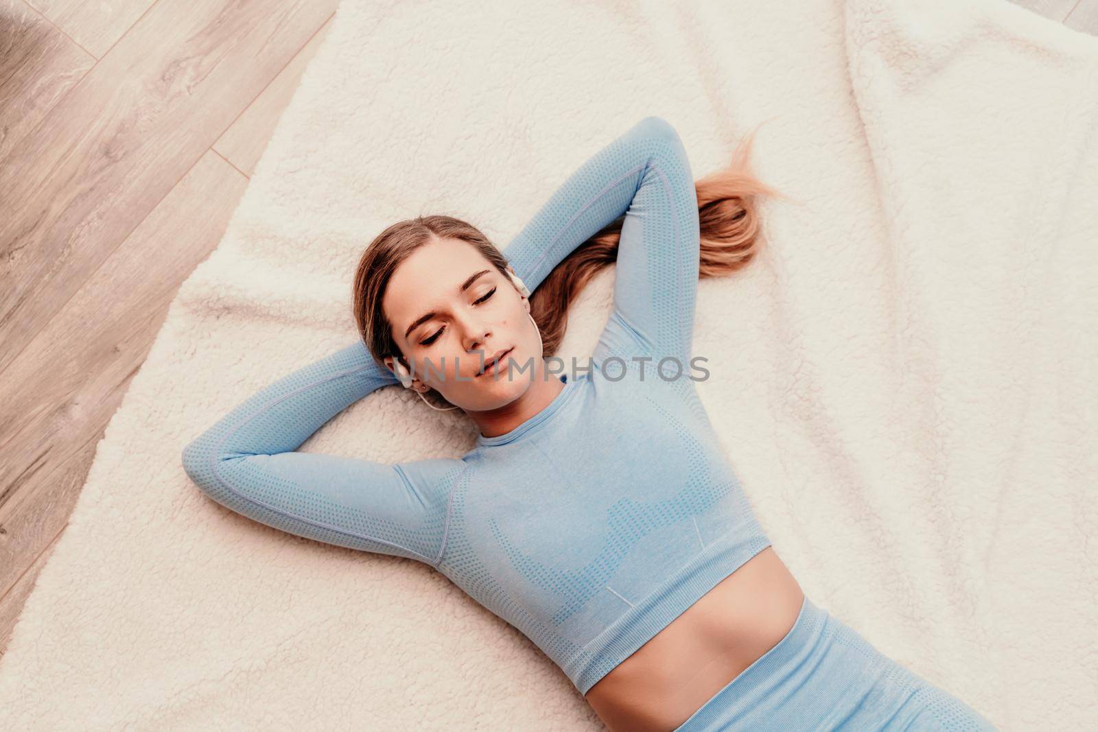 Top view portrait of relaxed woman listening to music with headphones lying on carpet at home. She is dressed in a blue tracksuit