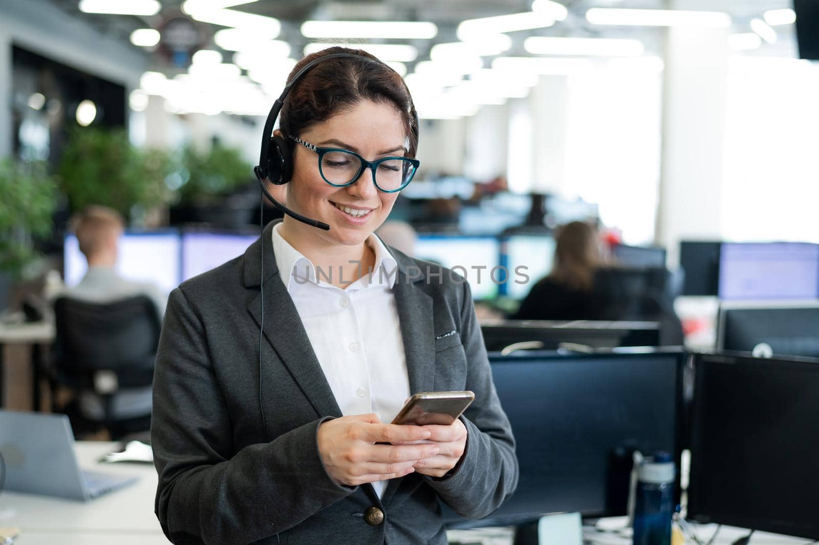 Beautiful caucasian woman in headset is holding a mobile phone while standing in open space office. Friendly female helpdesk operator browsing the screen of a smartphone in the workplace