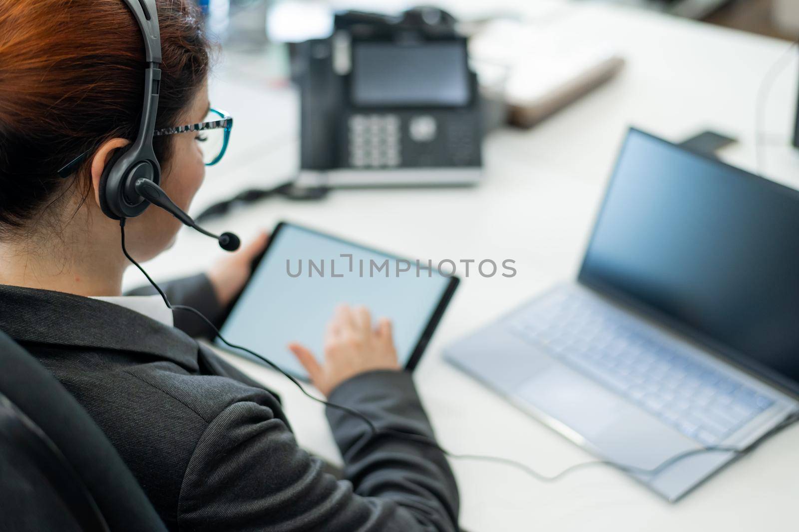 A business woman with a headset holds a digital tablet while sitting at a desk.