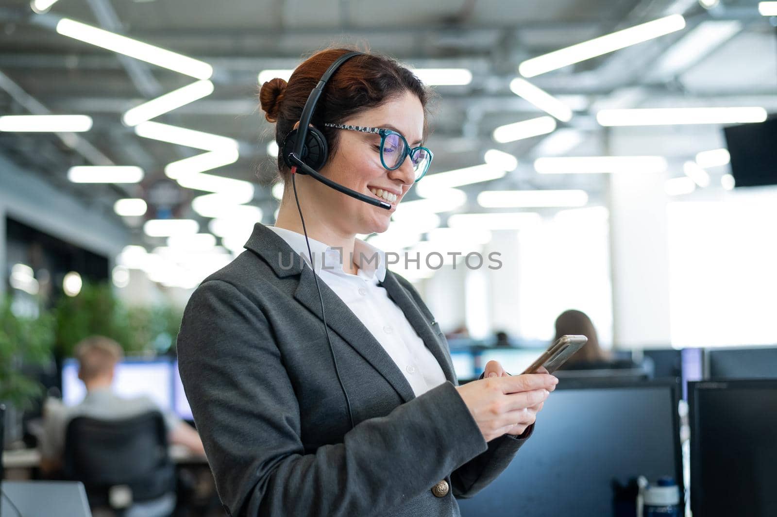 Beautiful caucasian woman in headset is holding a mobile phone while standing in open space office. Friendly female helpdesk operator browsing the screen of a smartphone in the workplace