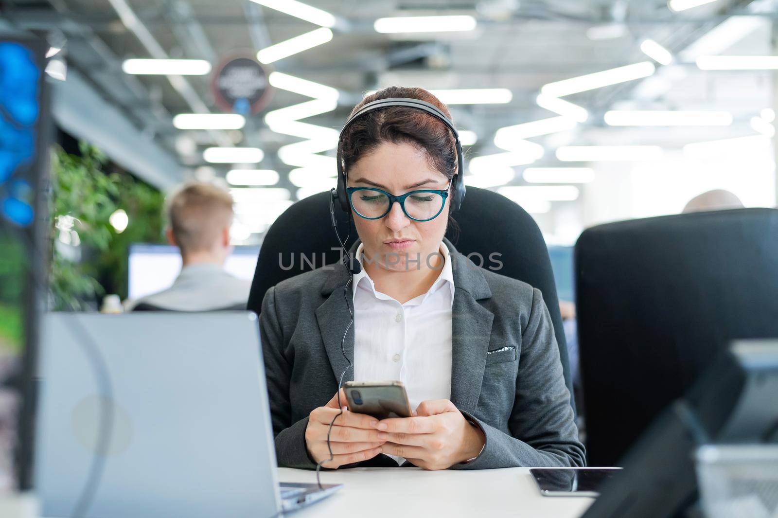 Business woman dressed in a headset is bored and uses a smartphone while sitting at a desk. Female manager is distracted from work by phone by mrwed54