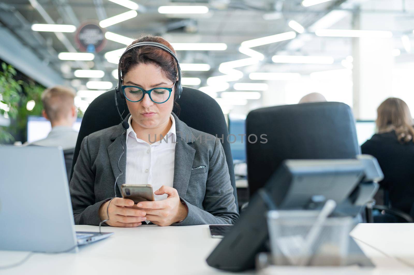 Business woman dressed in a headset is bored and uses a smartphone while sitting at a desk. Female manager is distracted from work by phone.