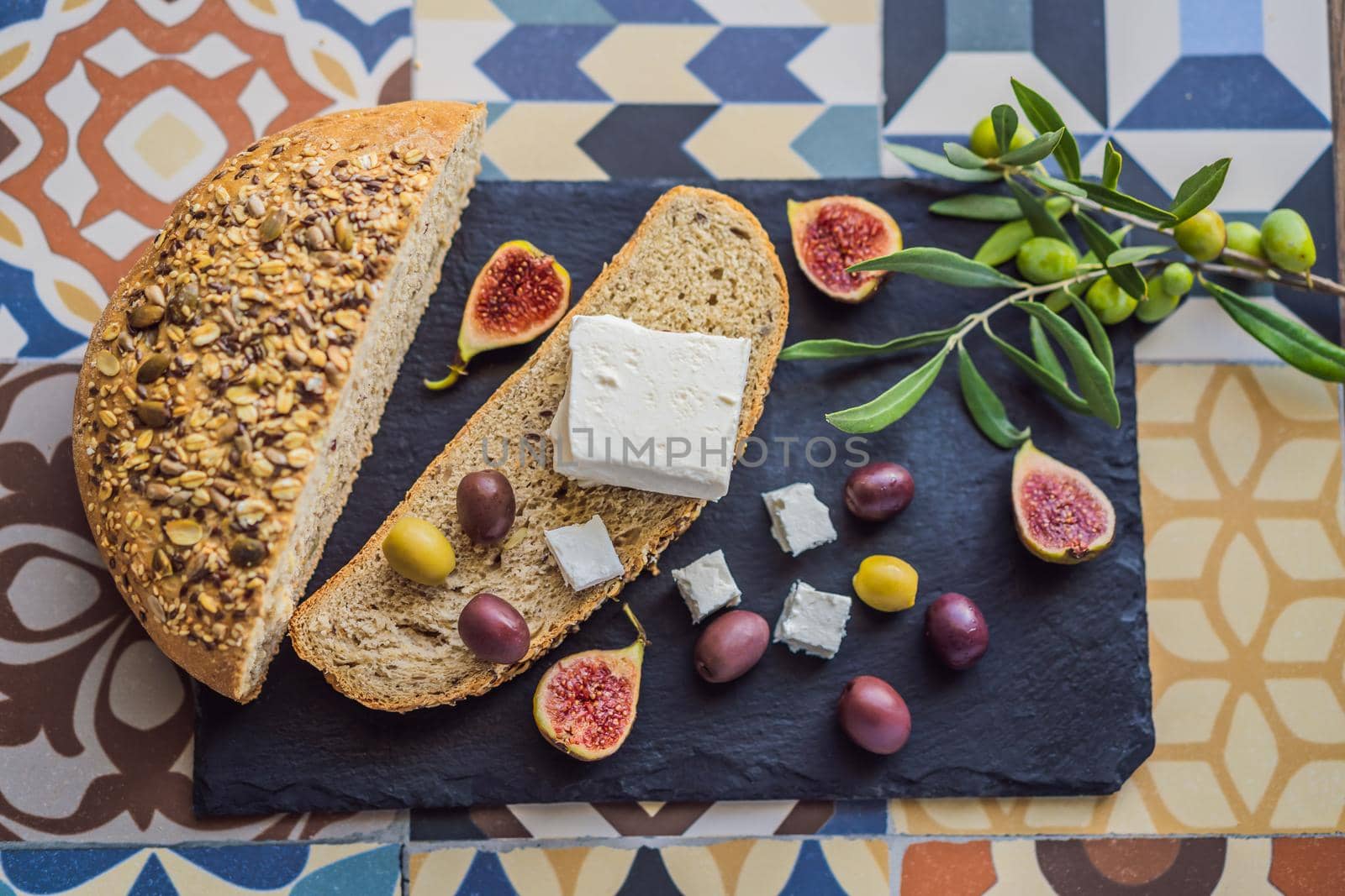 Green and black olives with loaf of fresh bread, feta cheese and young olives branch on olive wood chopping board over dark background.